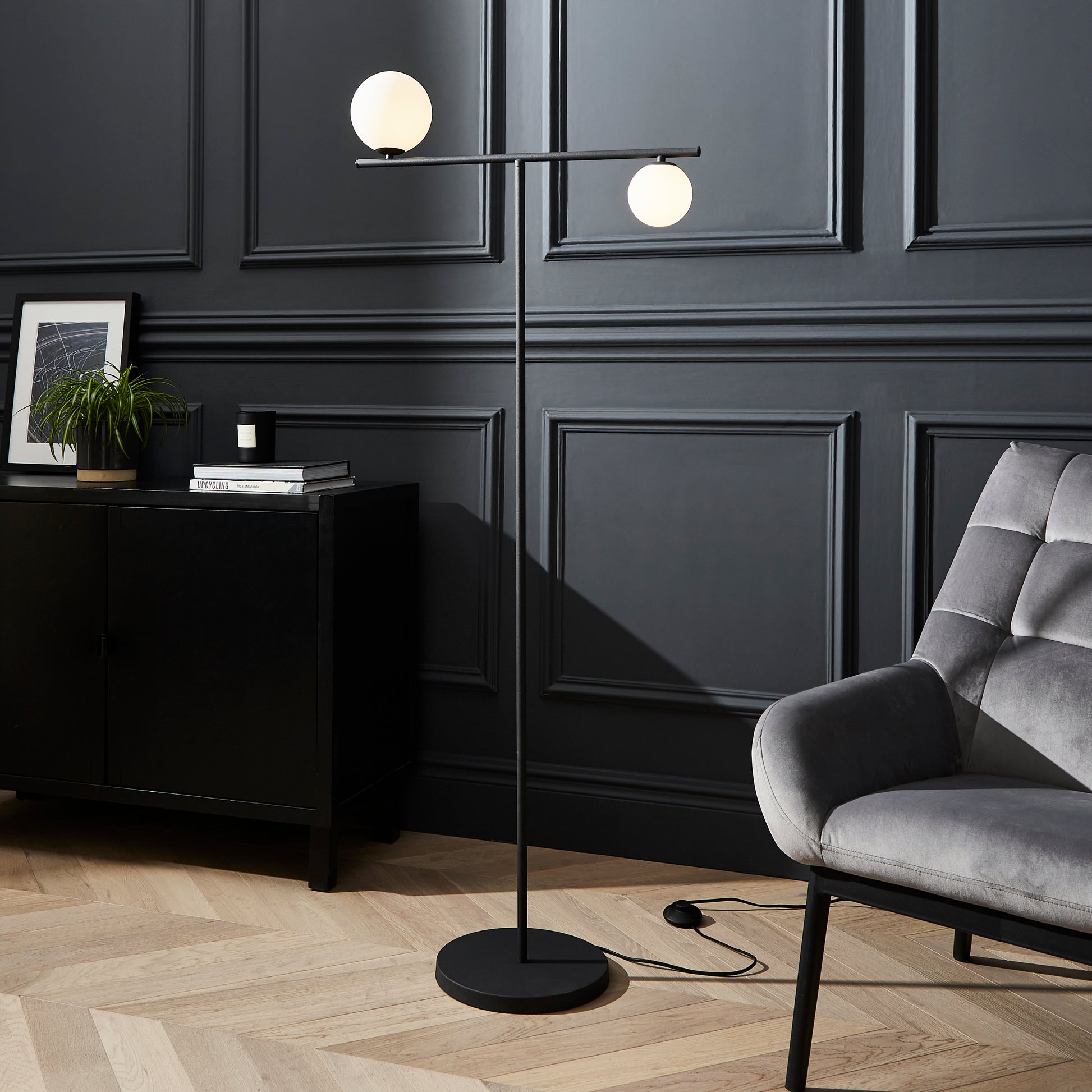 Modern floor lamp with two spherical lights in a room with dark paneled walls and a gray armchair.