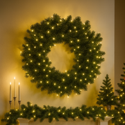 Decorative wreath with lights on a wall above a fireplace with candles and more greenery.
