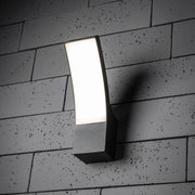 A modern curved LED wall light with a dark grey anthracite finish and an opal diffuser installed on a textured wall surface.