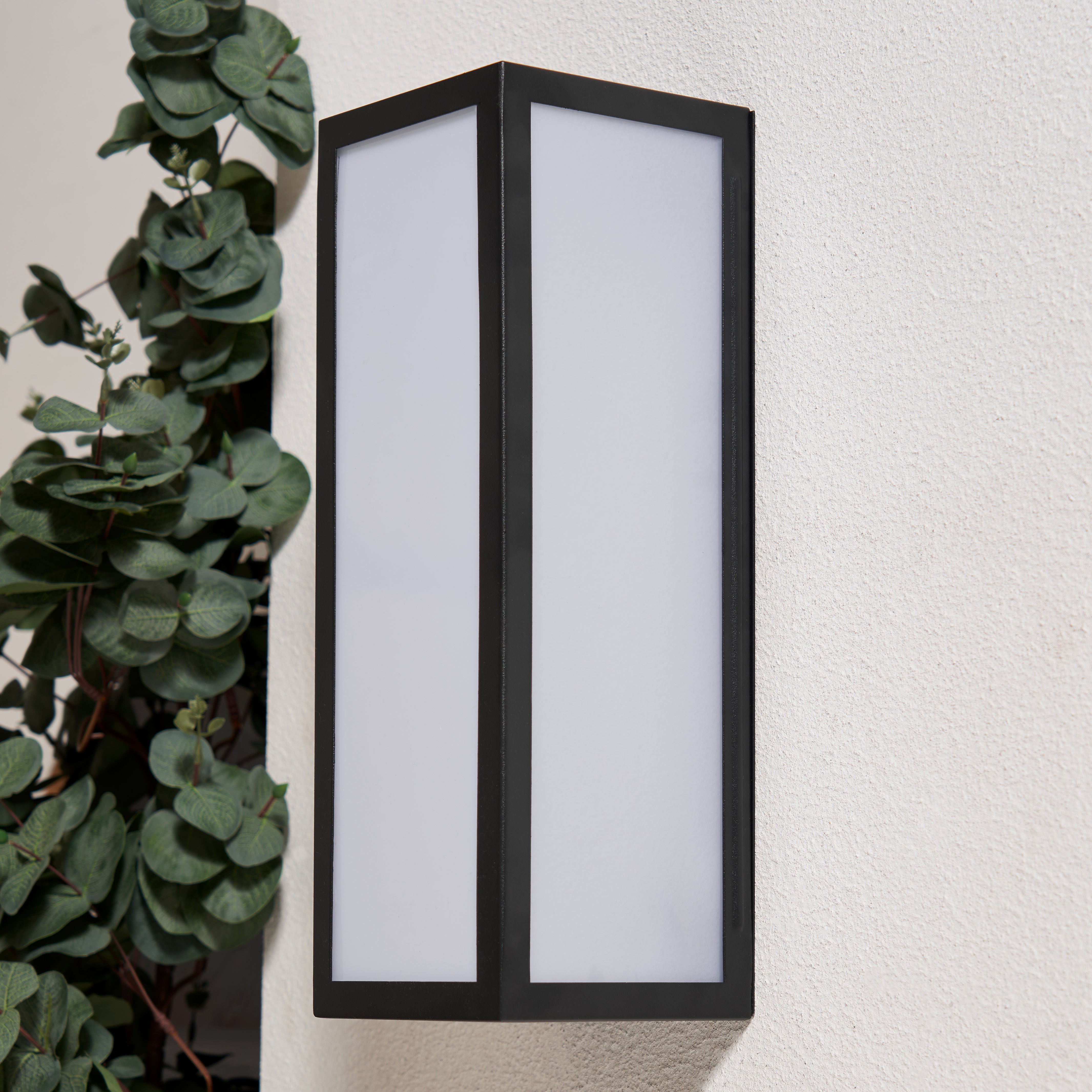 Black outdoor wall light fixture on a white wall with greenery to the left.