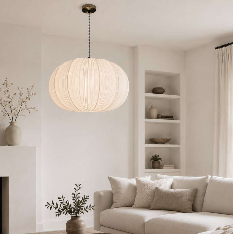 A modern living room featuring a large, spherical pleated pendant light hanging over a light wood coffee table. The room is styled in neutral tones with a cream sectional sofa, a white fireplace, and built-in shelves in the background.