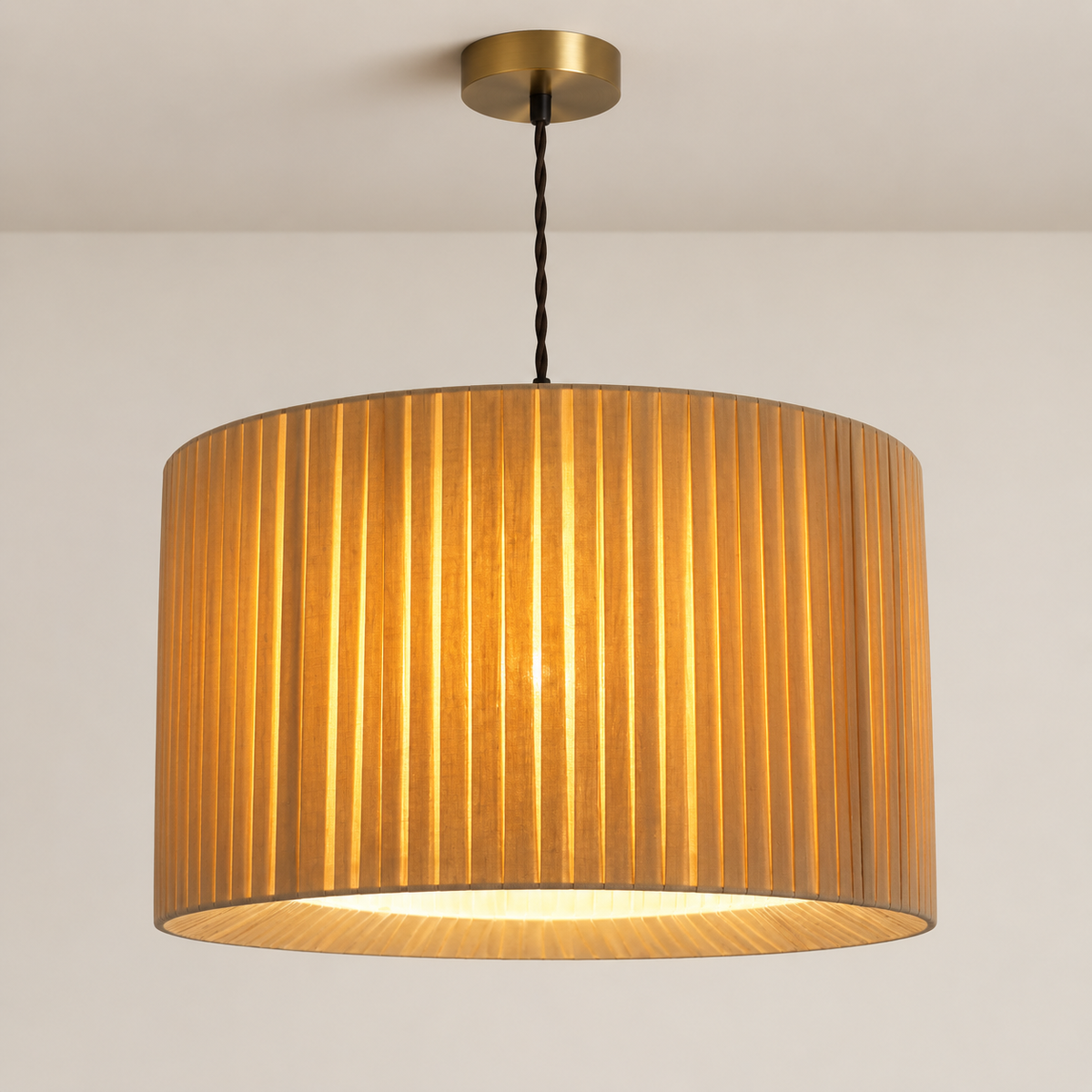 A modern drum pendant light with a pleated fabric shade in a warm golden-brown color, suspended by a black twisted cord from a round brass ceiling canopy.