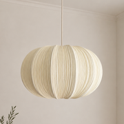 A cream-colored pleated paper pendant light hanging from a brass ceiling fixture in a minimalist, warm-toned room with a wooden table and vase.