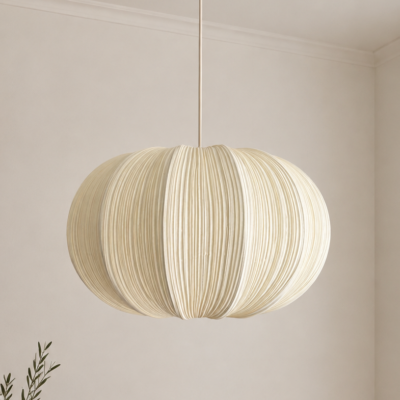 A cream-colored pleated paper pendant light hanging from a brass ceiling fixture in a minimalist, warm-toned room with a wooden table and vase.