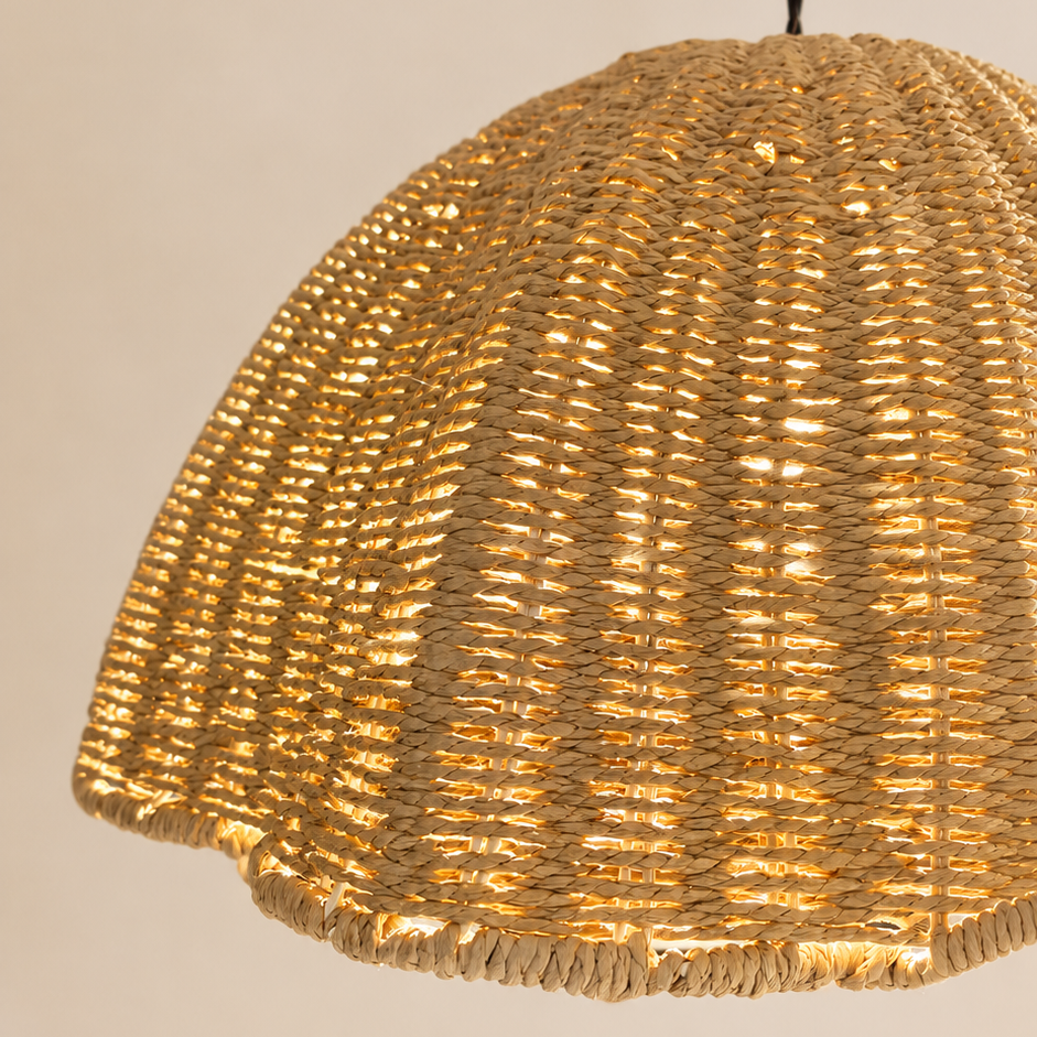 A close-up of a woven dome pendant light shade made from natural fiber, glowing warm yellow from within, hanging against a plain beige wall.