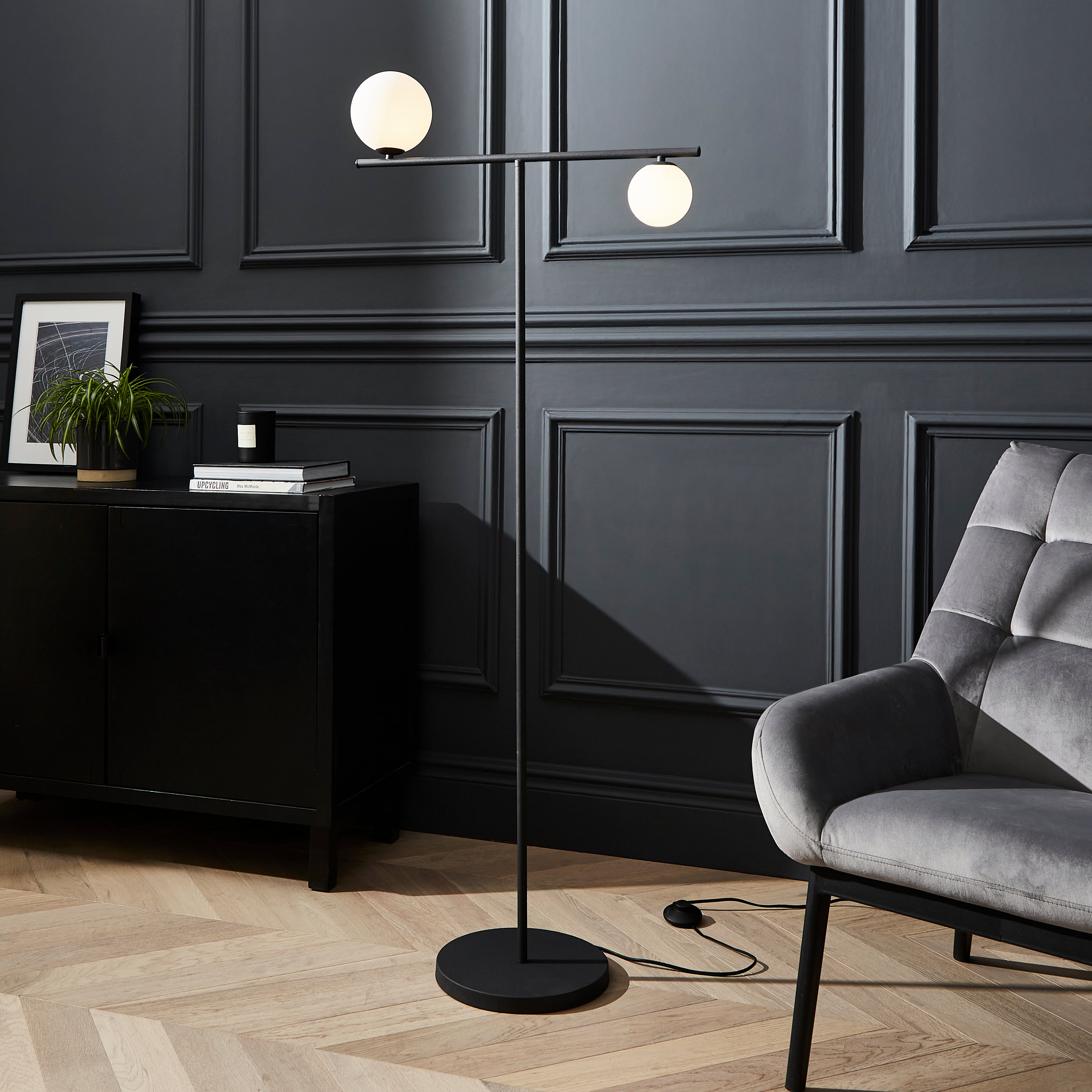 Modern floor lamp with two spherical lights in a room with dark paneled walls and a gray armchair.