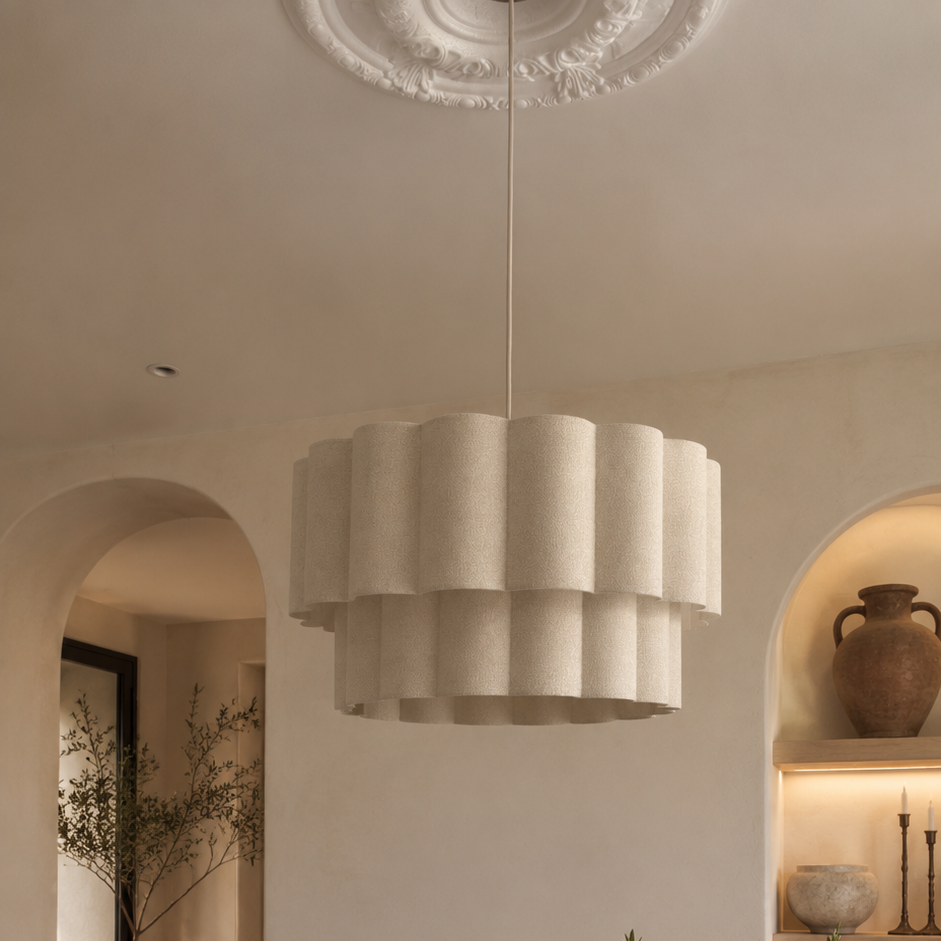 A modern, tiered pleated pendant light in a warm off-white or cream color hangs over a round wooden dining table with woven chairs in a minimalist, warm-toned dining room.