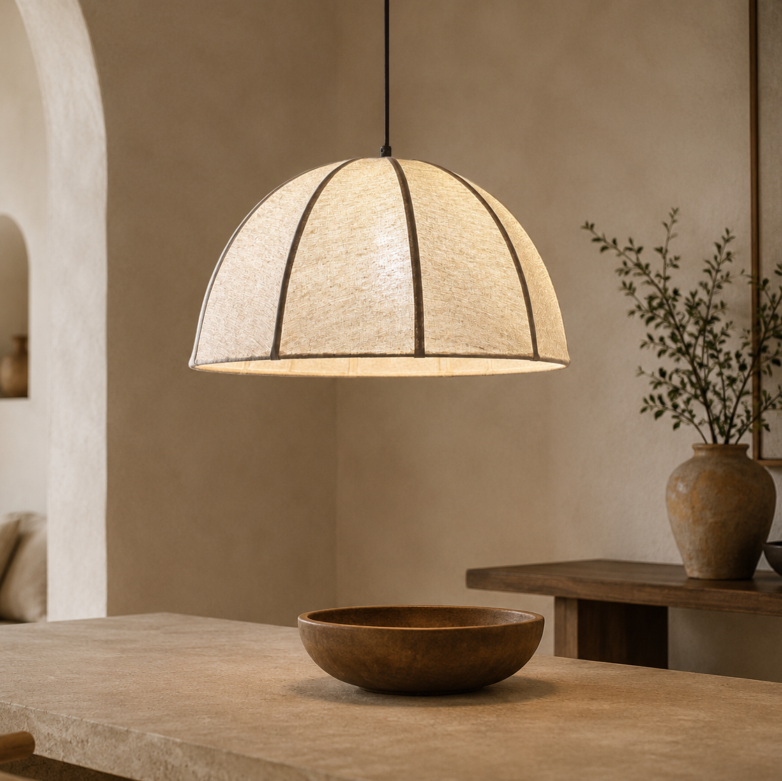 A dome-shaped pendant light with a textured beige woven shade and dark metal ribs, hanging in a warm, rustic room with a wooden bowl on a table below.