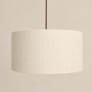 A drum-shaped pendant light shade made of textured cream linen fabric, suspended by a thin dark brown cord against a solid beige background.