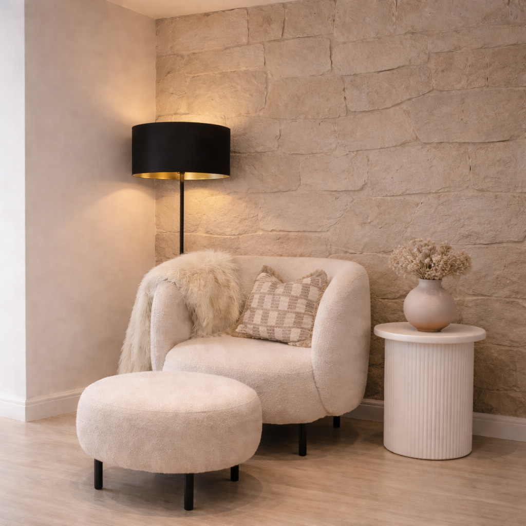 Modern living room with a beige armchair, ottoman, and side table against a stone wall.