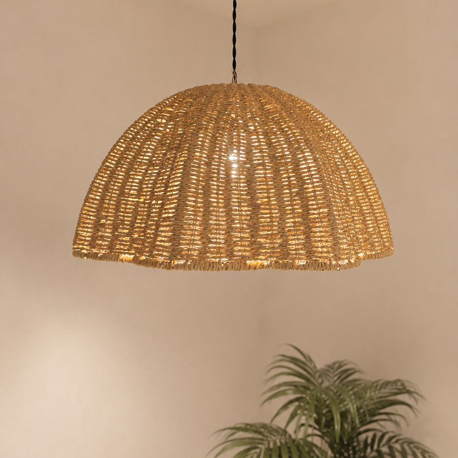 A dome-shaped woven pendant light in a natural wicker or rattan finish, hanging from a black cord and brass ceiling canopy. The light is turned on, casting a warm glow and a circular light pattern on the white ceiling.