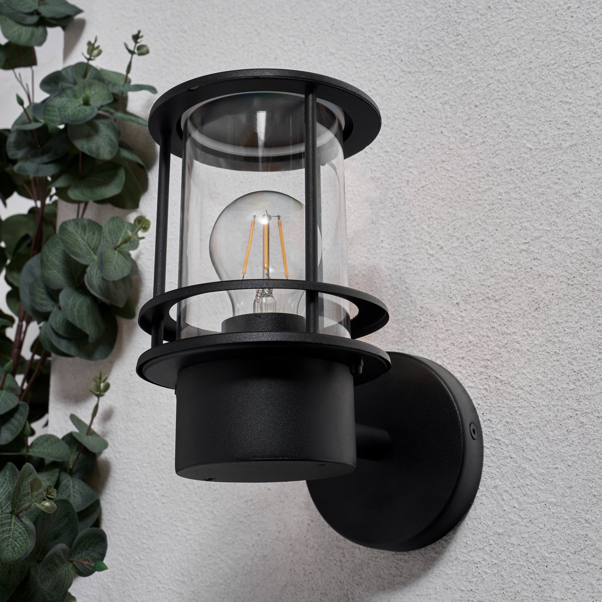 Black outdoor wall light fixture on a gray wall with greenery to the left.