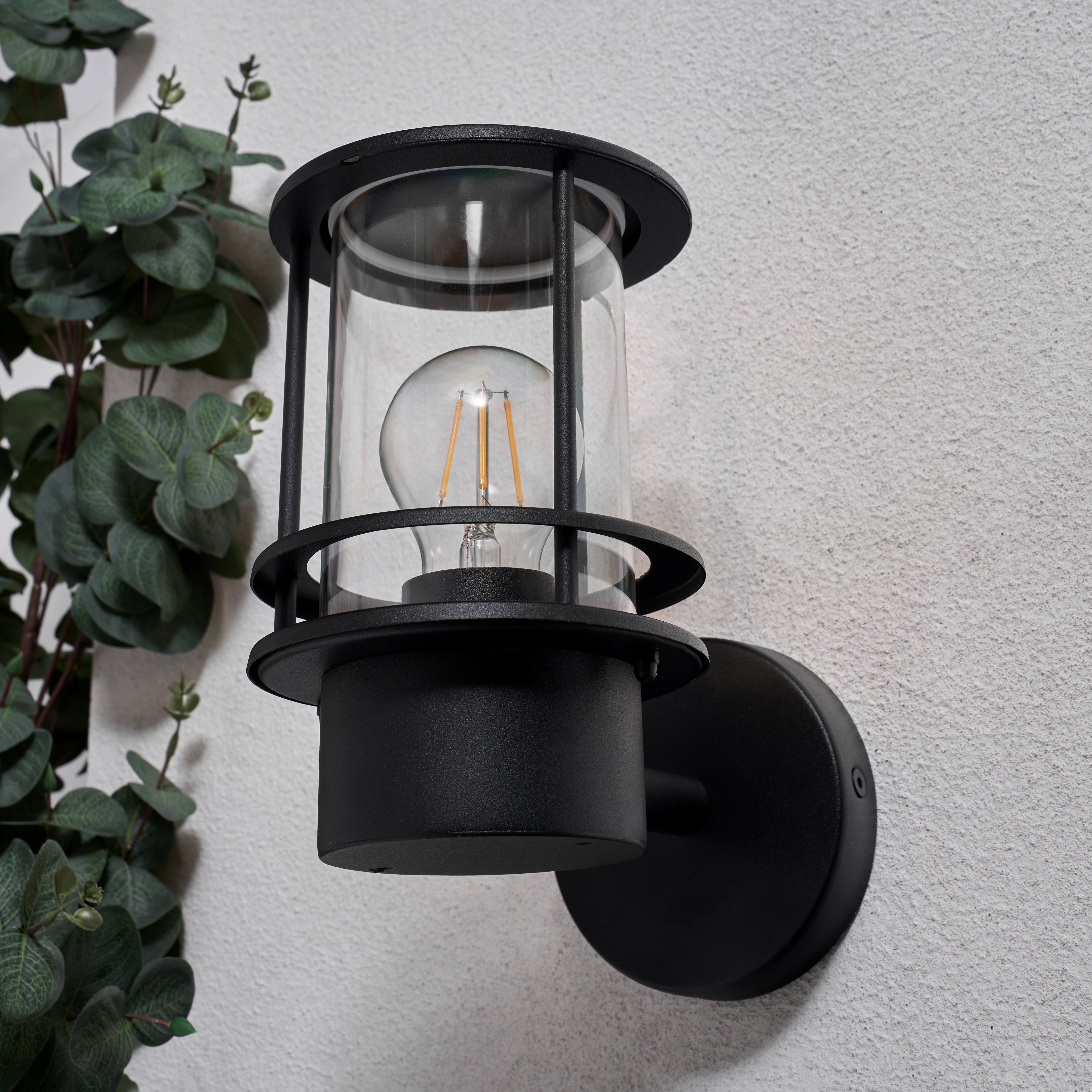 Black outdoor wall light fixture on a gray wall with greenery to the left.