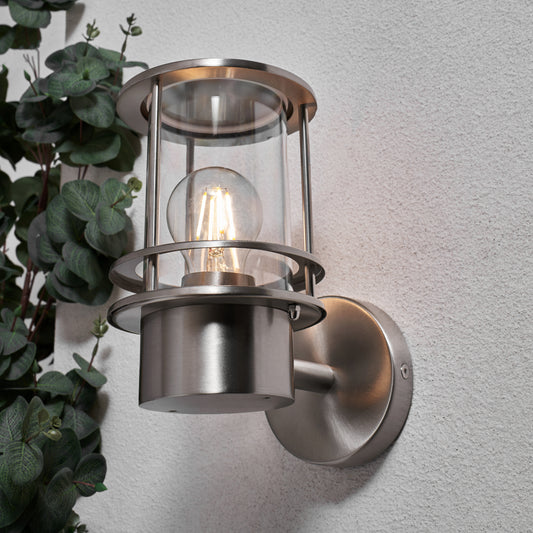 Metallic outdoor wall light fixture on a gray wall with a plant to the left.