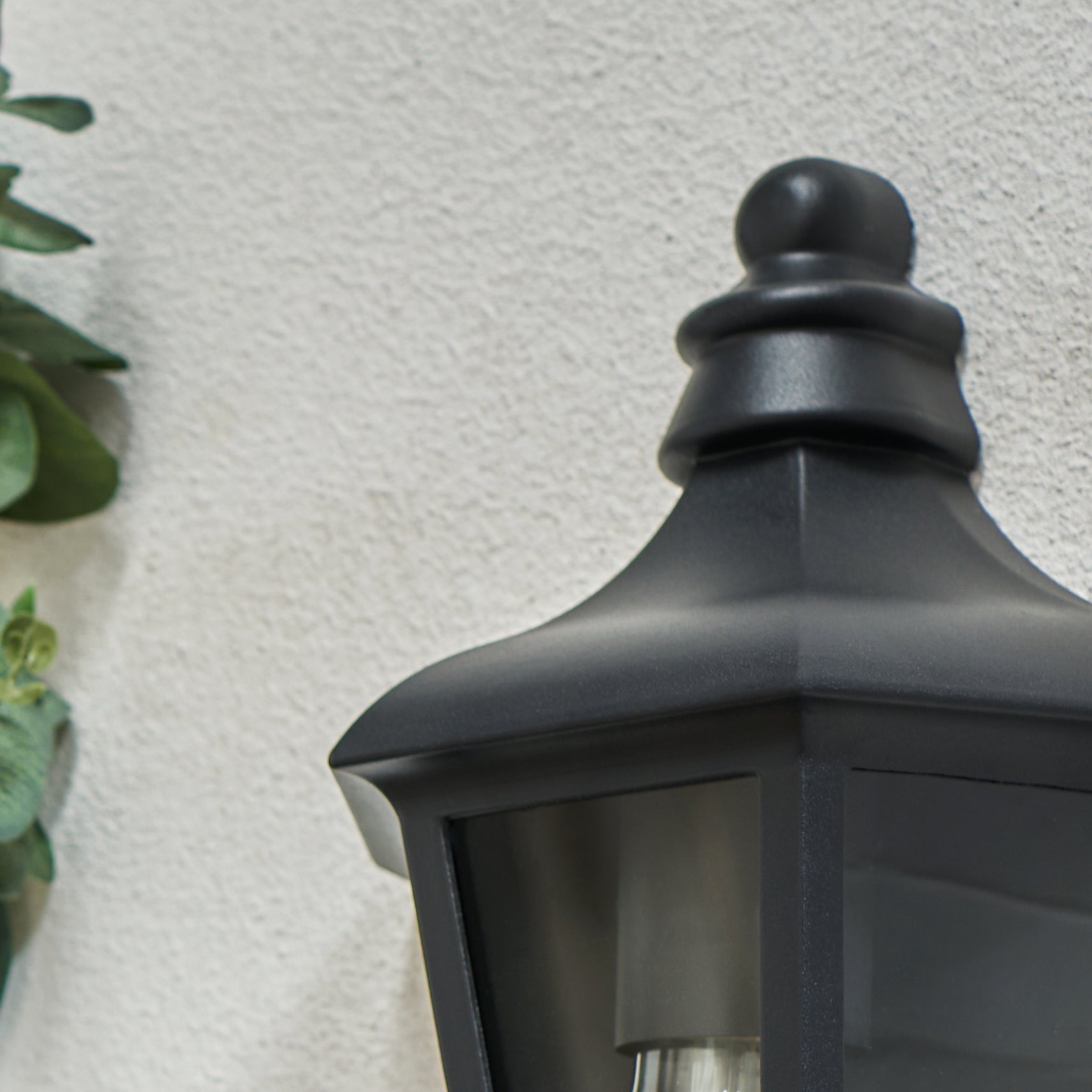Black outdoor wall light fixture on a textured white wall with a plant in the corner.