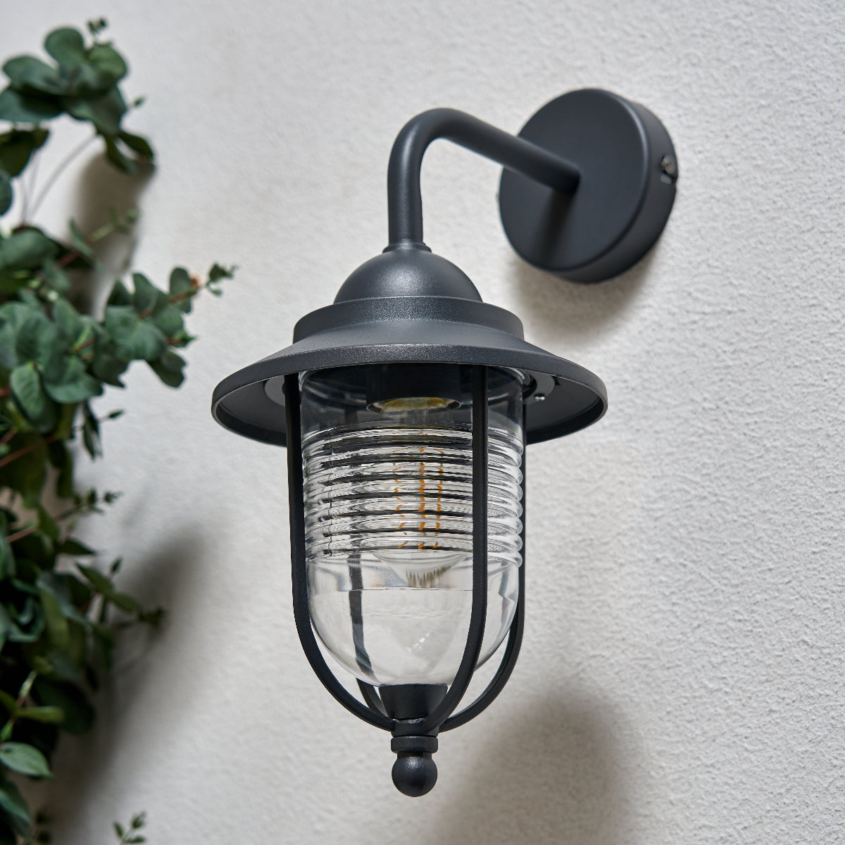 Black outdoor wall light fixture on a light gray wall with a plant to the left.