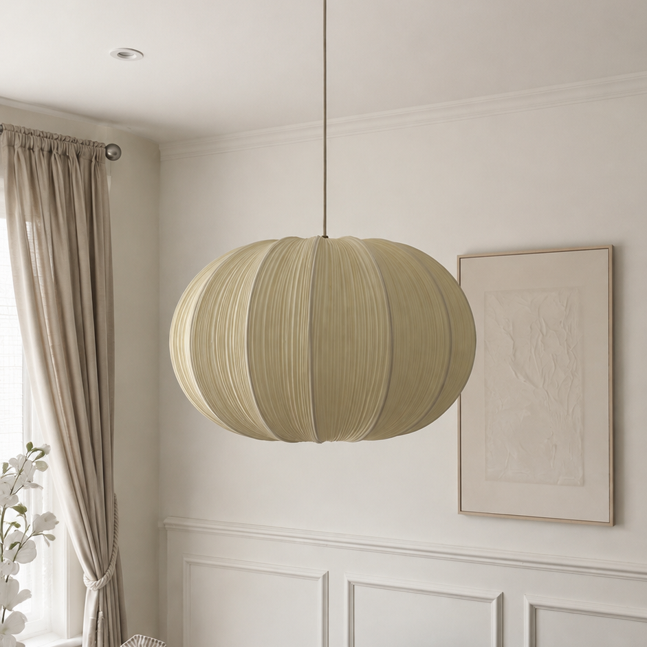 A beige pleated paper lantern pendant light hanging from a brass fixture in a white ceiling, set in a beautifully styled room with white paneled walls, beige curtains, and a black radiator.