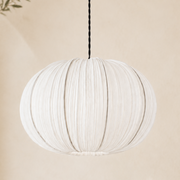 A white pleated fabric pendant light hangs from a black cord against a warm beige wall, with soft shadows and a small leafy branch visible at the left edge.