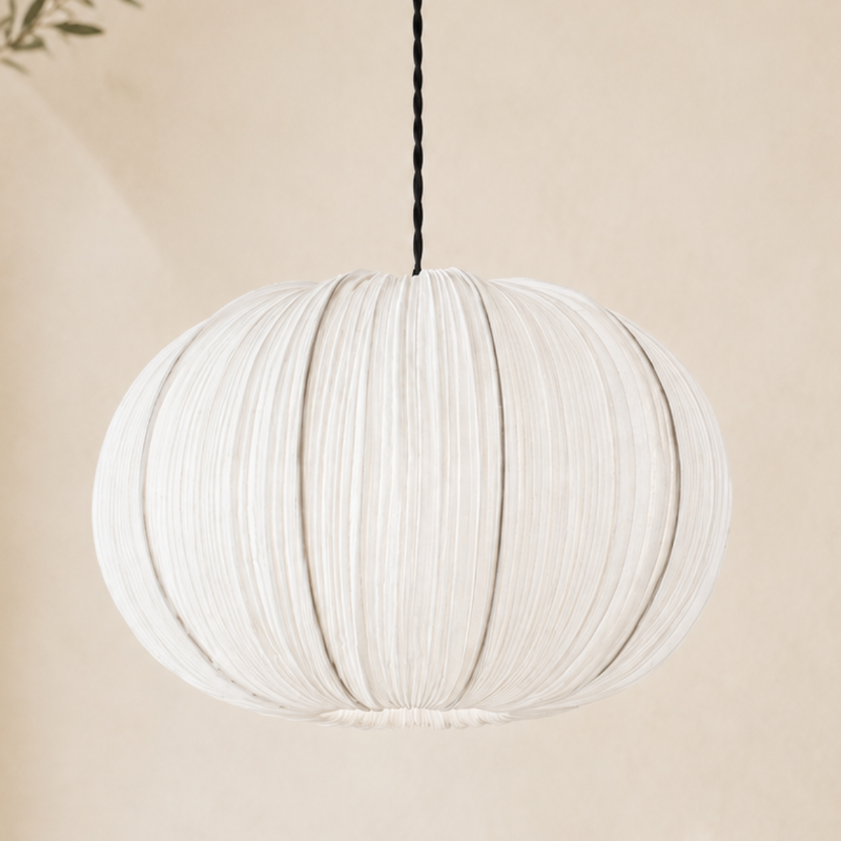 A white pleated fabric pendant light hangs from a black cord against a warm beige wall, with soft shadows and a small leafy branch visible at the left edge.