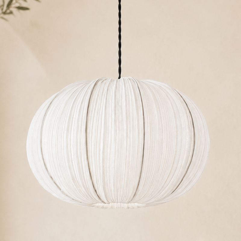 A white pleated fabric pendant light hangs from a black cord against a warm beige wall, with soft shadows and a small leafy branch visible at the left edge.