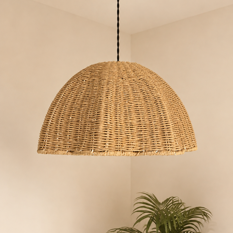 A dome-shaped woven rattan pendant light hanging from a brass ceiling canopy in a minimalist room corner with a potted palm plant below.