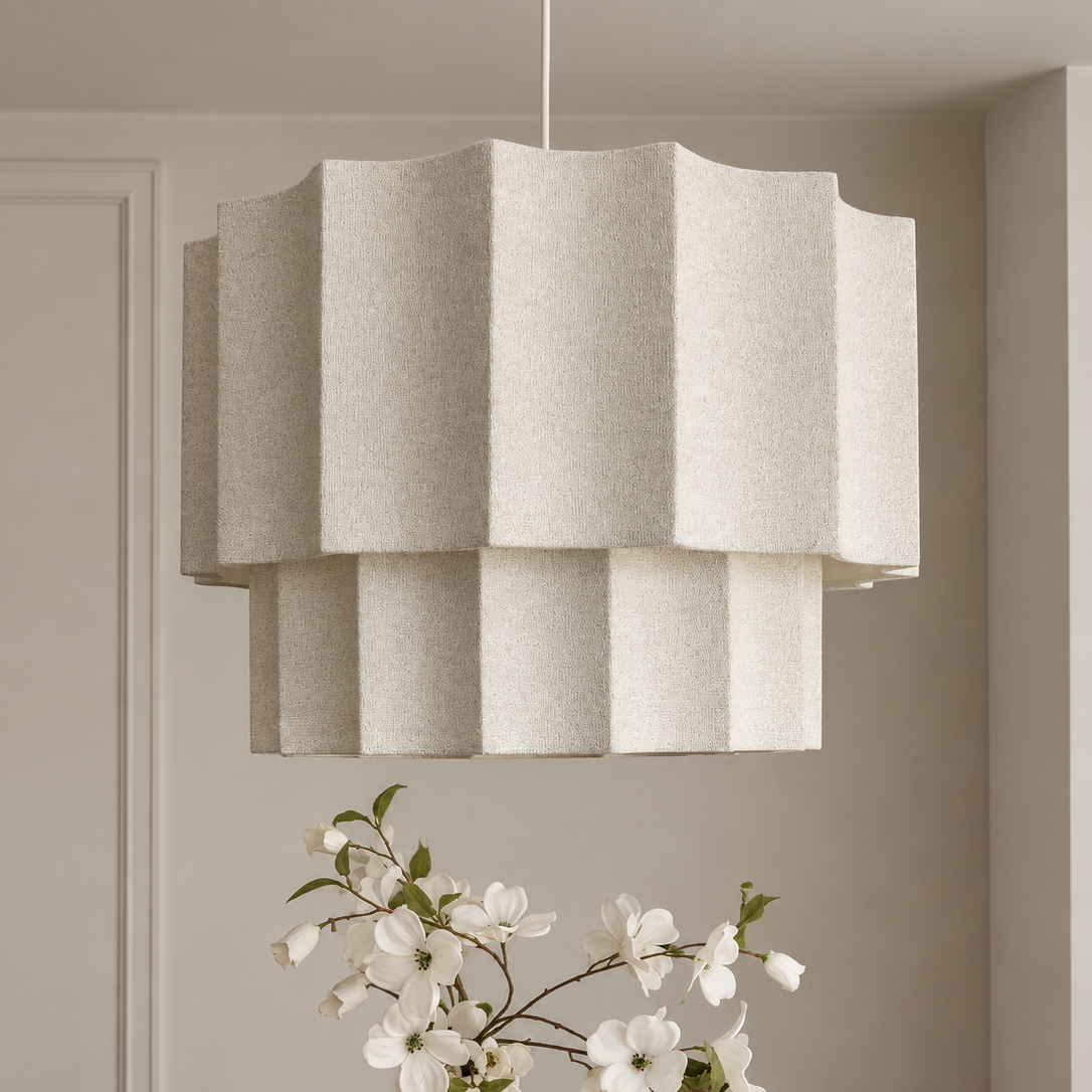 A modern multi-tiered pendant light with a textured off-white felt shade hangs over a wooden dining table. A white ceramic vase with white blossoms sits on the table in a softly lit, neutral-toned room.
