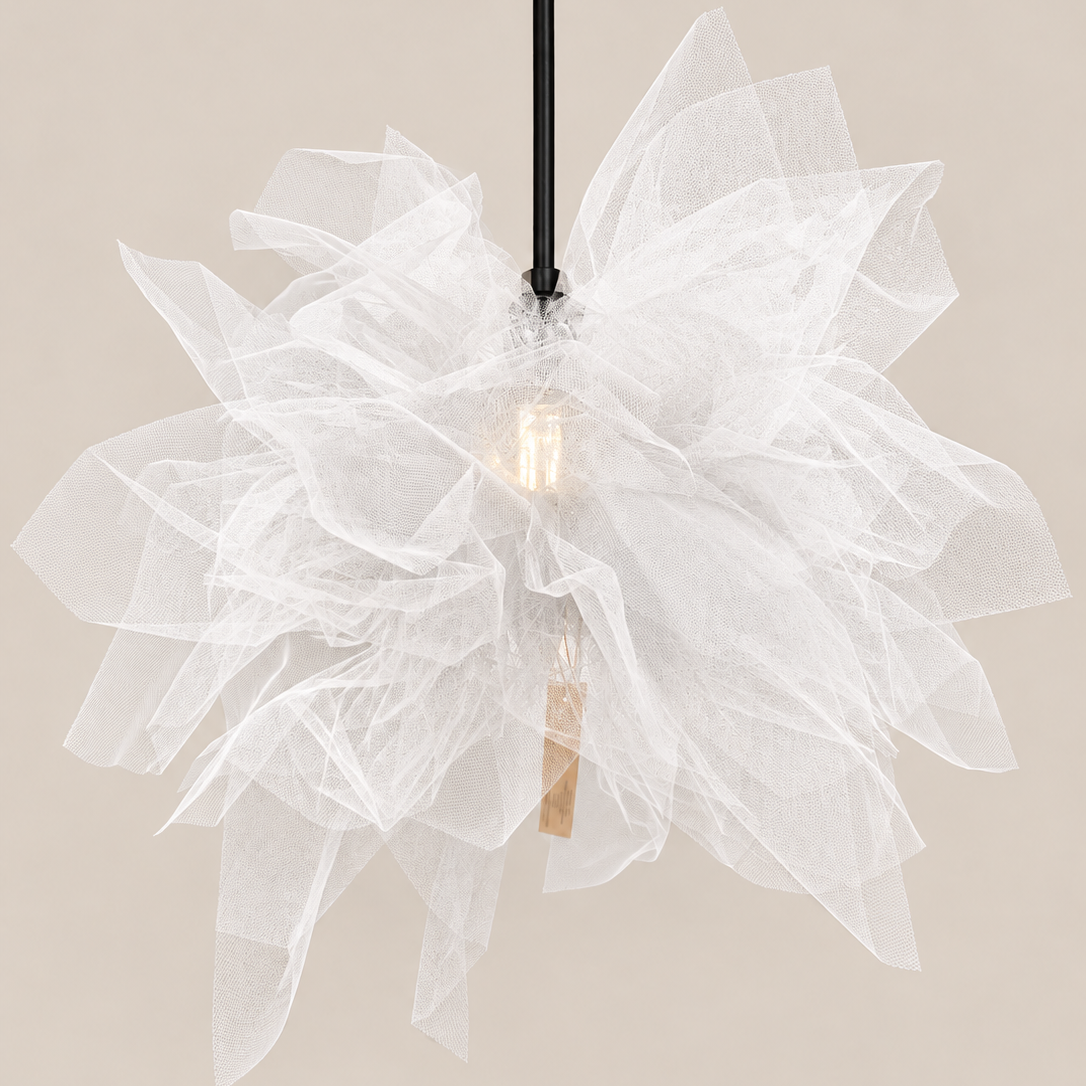A modern pendant light with a large, layered white tulle flower head, a black hanging cord, and a visible warm-toned Edison bulb inside, set against a solid beige background.