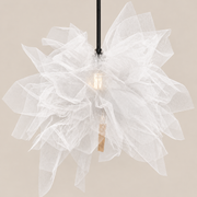 A modern pendant light with a large, layered white tulle flower head, a black hanging cord, and a visible warm-toned Edison bulb inside, set against a solid beige background.