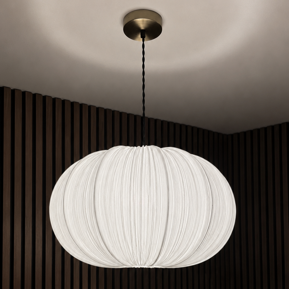 A modern white pleated pendant light hanging from a brass ceiling canopy against a dark brown slatted wall.