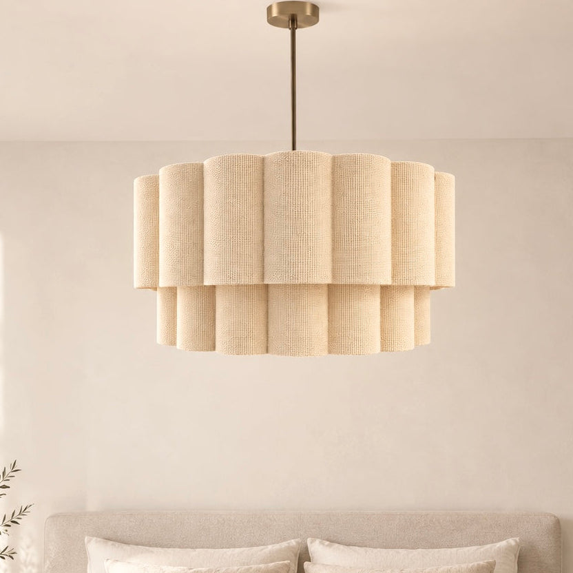A modern tiered drum pendant light with a textured beige linen shade hangs over a neatly made bed in a softly lit, minimalist bedroom.