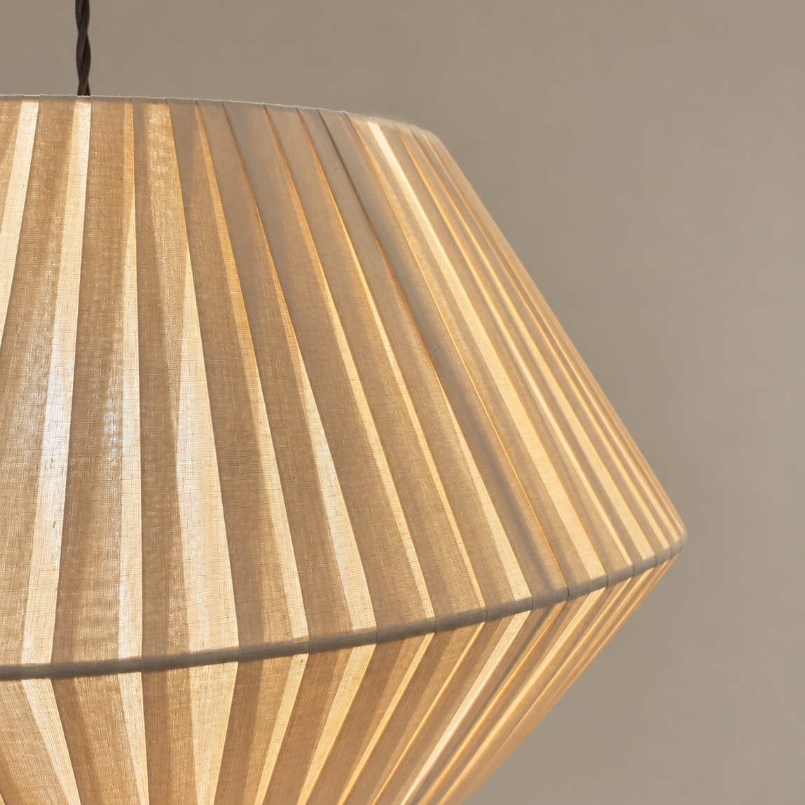 A close-up of a pleated fabric lampshade in a warm beige or light wood tone, glowing softly against a plain neutral background.