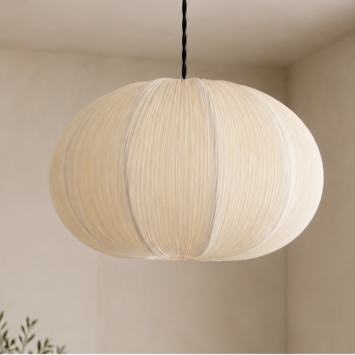 A modern spherical pendant light with pleated cream fabric and a brass ceiling canopy, hanging in a minimalist beige-toned room.