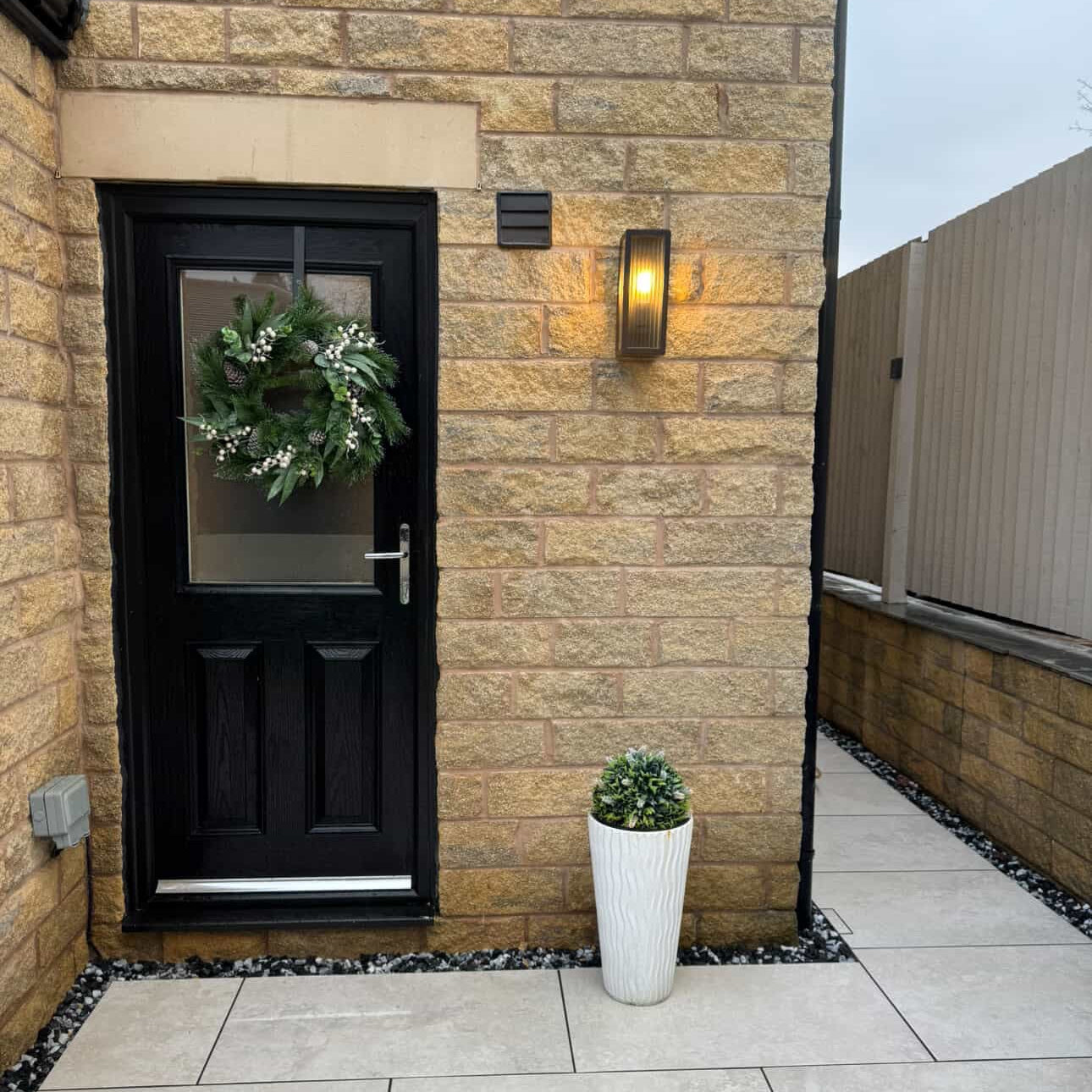 Black door with wreath on a stone wall, light fixture, and potted plant.