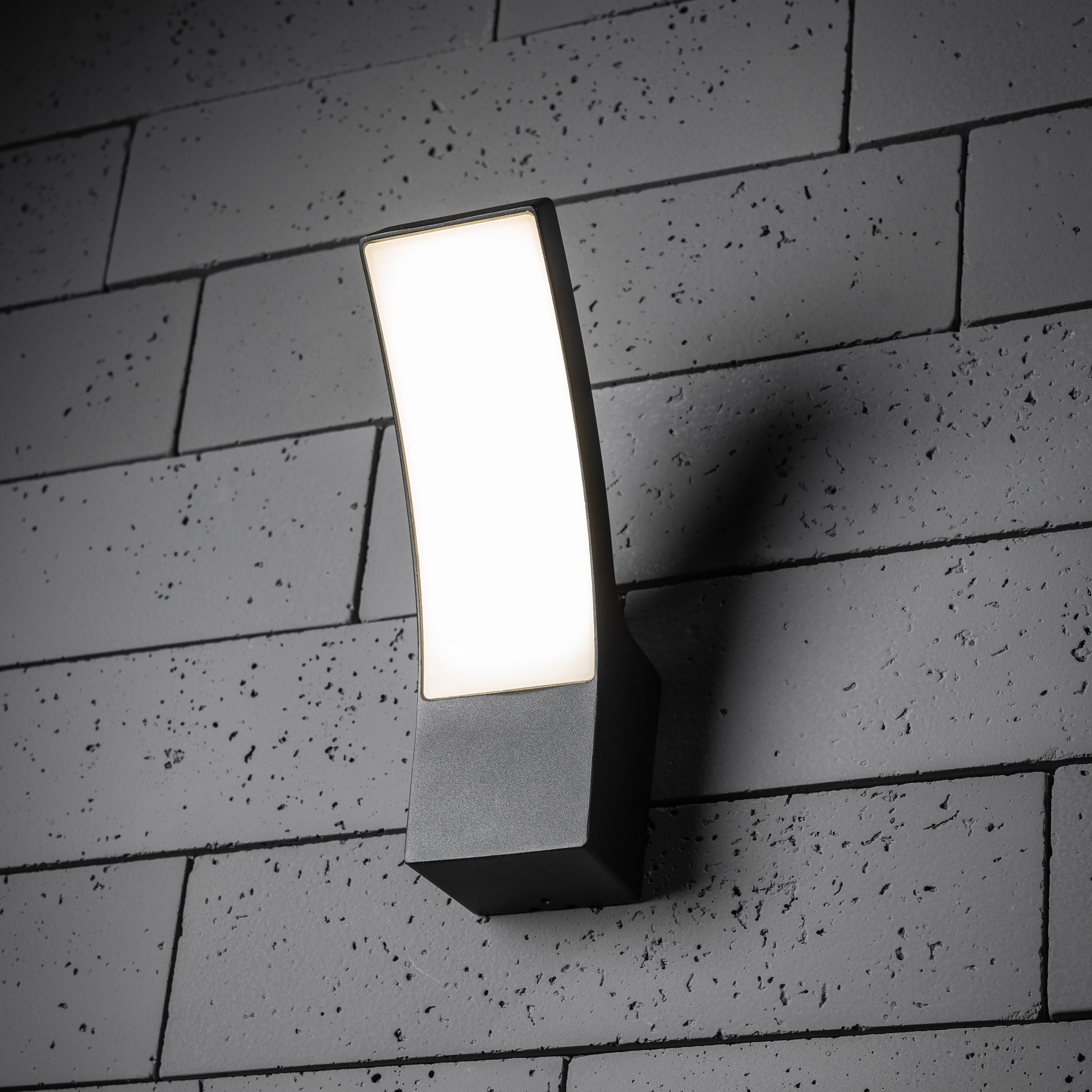 A modern curved LED wall light with a dark grey anthracite finish and an opal diffuser installed on a textured wall surface.