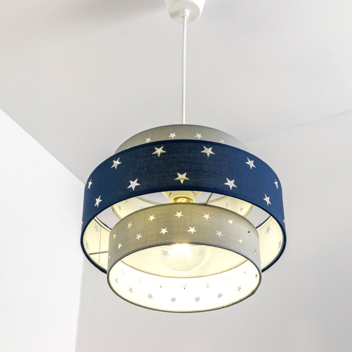 Our grey and navy two tiered star shade features a mixture of large and small sweet stars cut out across a grey and navy background. When your lamp is turned on it will shine out through these cut-outs, mimicking a beautiful night sky. This lampshade is made with soft-touch fabric for a little bit of luxury.