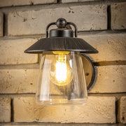 A black and bronze wall lantern with a glass shade illuminated, mounted on a brick wall.