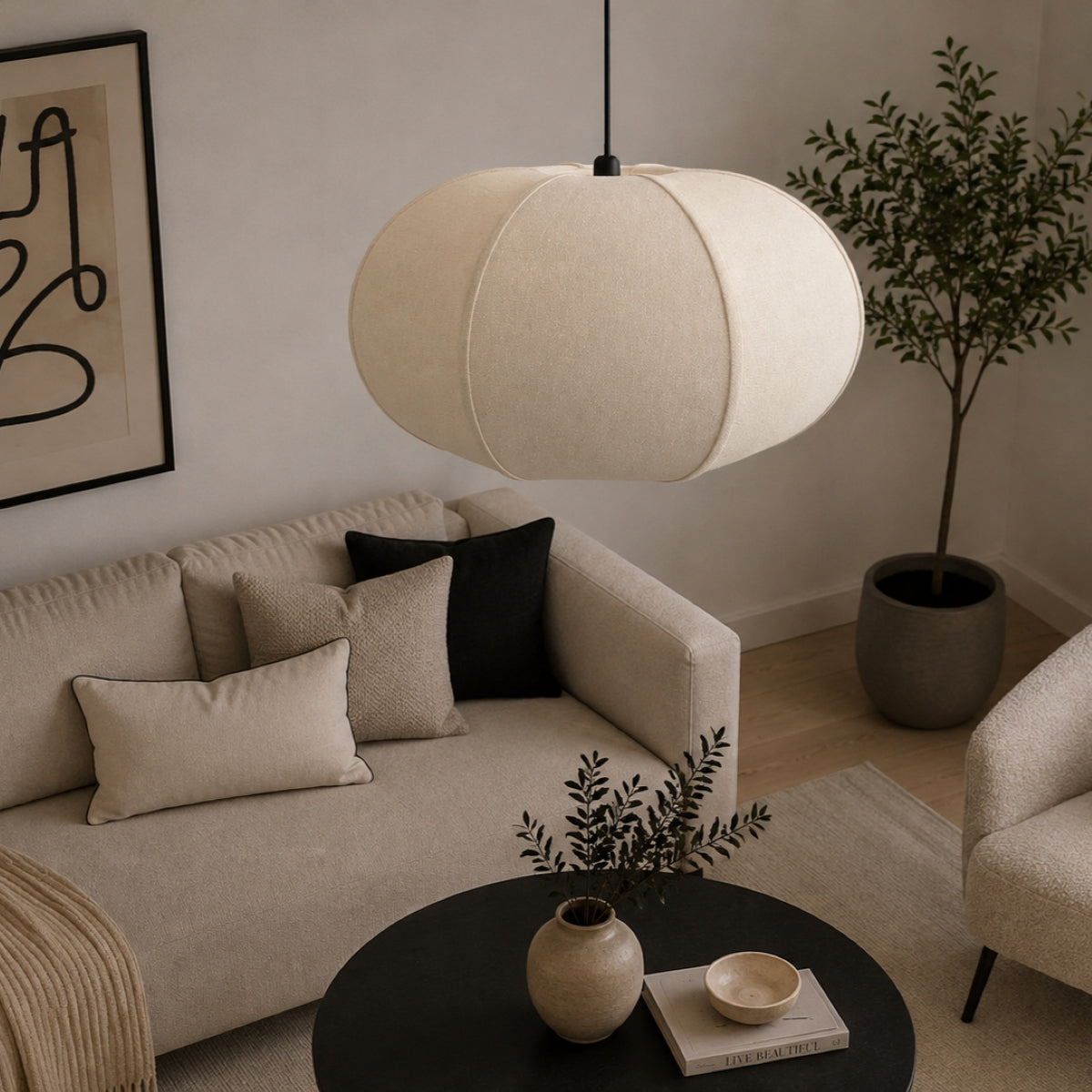 A modern pendant light with a large, spherical, fabric-covered shade in a warm cream or off-white color, hanging in a styled living room over a black coffee table.