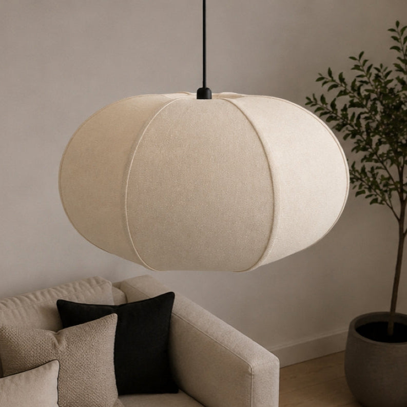 A modern pendant light with a large, spherical, fabric-covered shade in a warm cream or off-white color, hanging in a styled living room over a black coffee table.