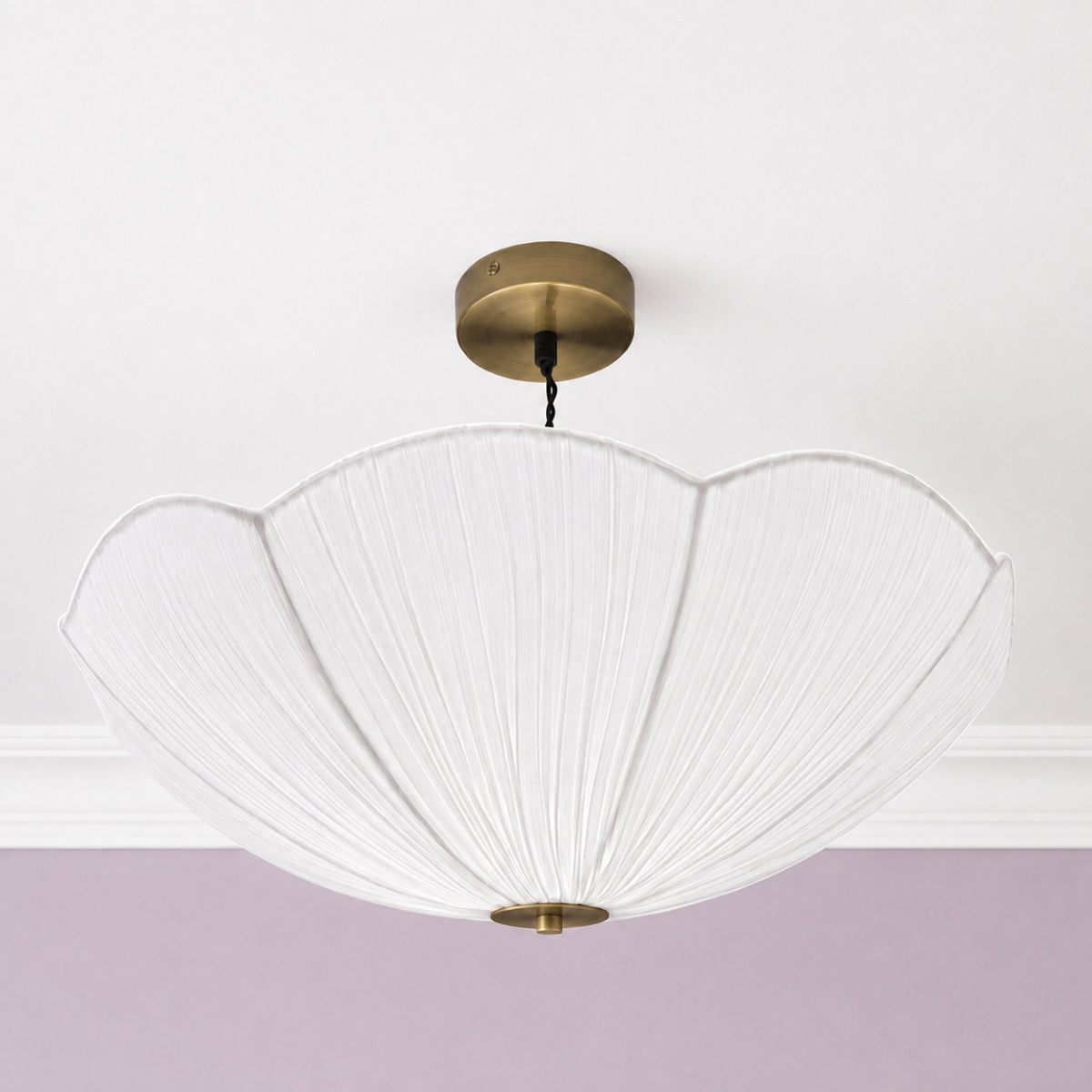 A modern ceiling light fixture with a large, pleated white fabric shade in a scalloped shape, suspended from a round brass ceiling canopy against a white ceiling and a lavender-painted wall.