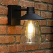 Black outdoor wall light fixture with clear glass shade on a brick wall.