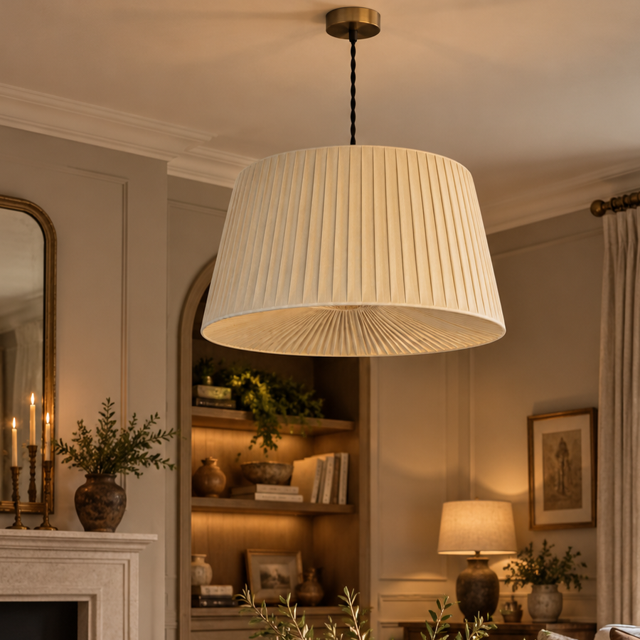 A large pleated drum pendant light in warm cream hangs over a rustic wooden coffee table in a cozy, elegantly decorated living room with a fireplace, sofa, and shelves.