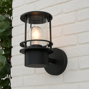 Black outdoor wall light fixture on a white brick wall with a plant to the left.