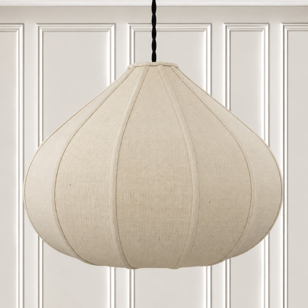 A large, teardrop-shaped pendant light with a textured beige linen shade, hanging from a black cord against a white paneled wall.