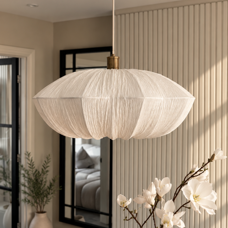 A modern pendant light with a textured, pleated white fabric shade hangs over a wooden dining table in a styled interior. The scene includes a white ceramic vase with white blossoms, a small white dish on a book, and a slatted wall background.
