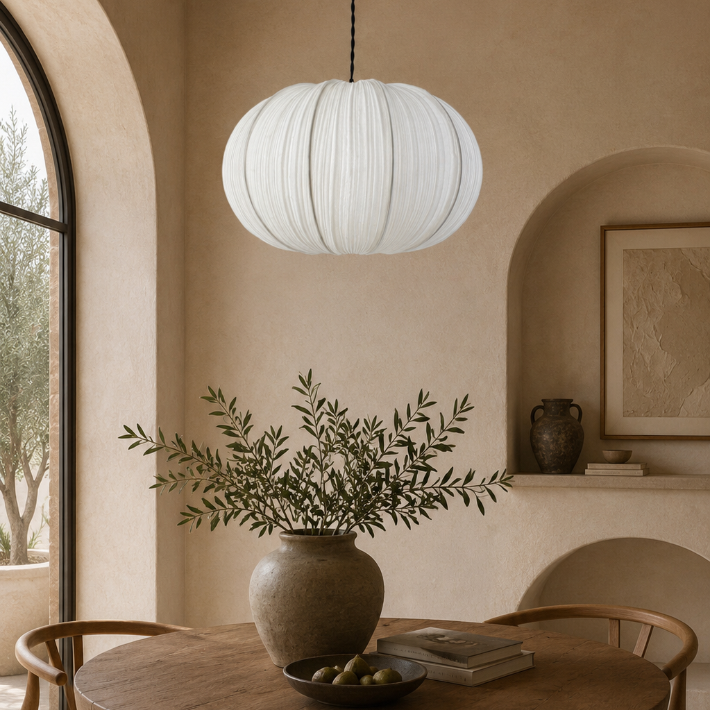 A white pleated paper lantern pendant light hangs over a round wooden dining table in a warm, rustic room with beige walls, wooden chairs, and a large ceramic vase with greenery.