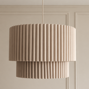 A modern two-tiered drum pendant light with a pleated fluted design in a warm beige tone, hanging against a matching beige paneled wall.