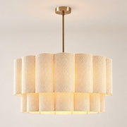 A modern drum pendant light with a tiered shade made of textured cream fabric panels, suspended by a slim brass rod from a matching round ceiling canopy.