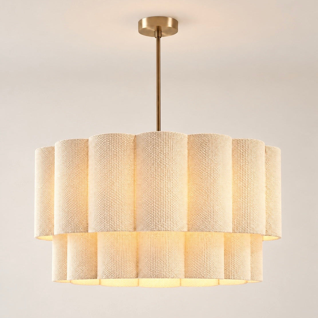 A modern drum pendant light with a tiered shade made of textured cream fabric panels, suspended by a slim brass rod from a matching round ceiling canopy.
