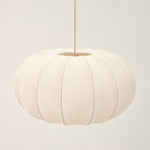 A modern pendant light with a ribbed, pumpkin-shaped white fabric shade and a brushed brass metal canopy and rod, shown hanging against a minimalist off-white wall.