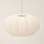 A modern pendant light with a ribbed, pumpkin-shaped white fabric shade and a brushed brass metal canopy and rod, shown hanging against a minimalist off-white wall.